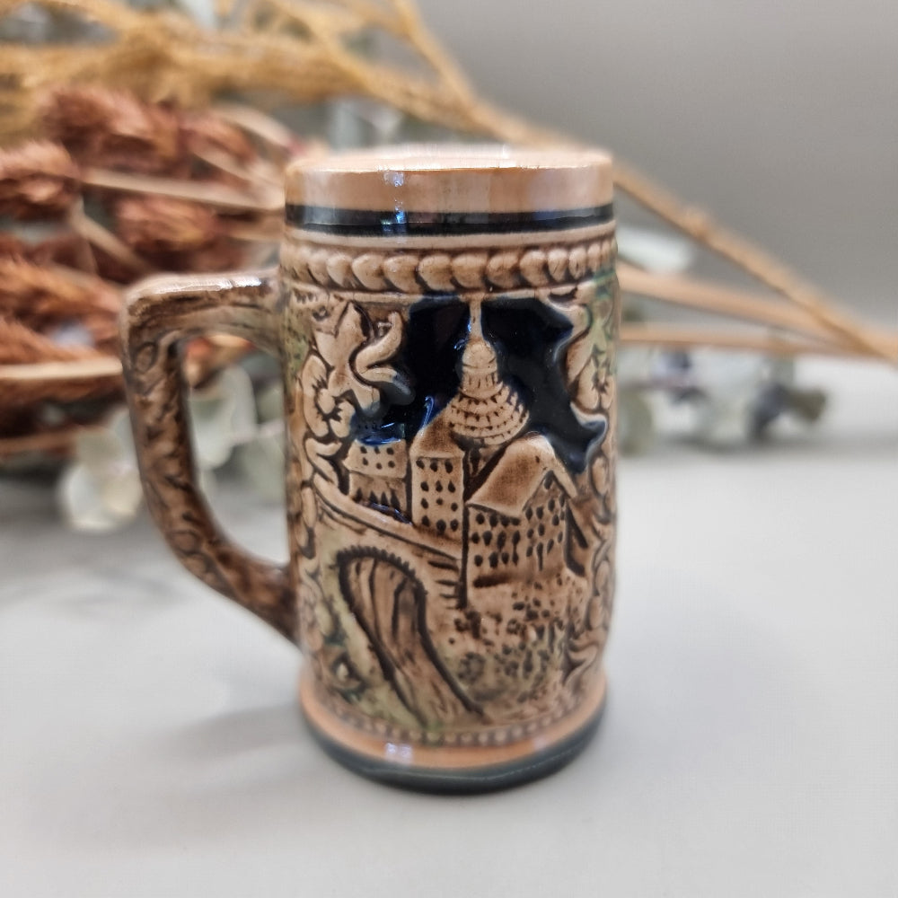 Miniature ceramic stein with embossed pastoral scene and castle design, hand-painted in earthy tones.