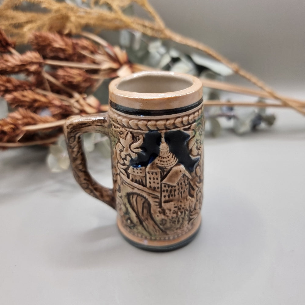 Miniature ceramic stein with embossed pastoral scene and castle design, hand-painted in earthy tones.