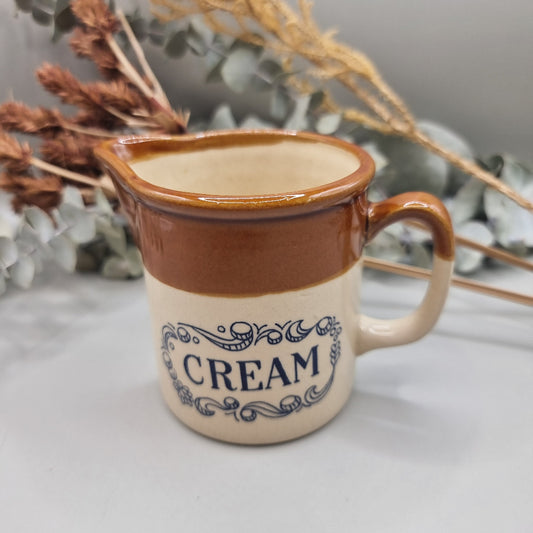 Small vintage stoneware cream jug with brown glaze and “CREAM” in blue lettering.