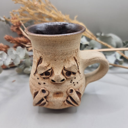 Stoneware face mug with grumpy sculpted expression and rustic brown glaze.