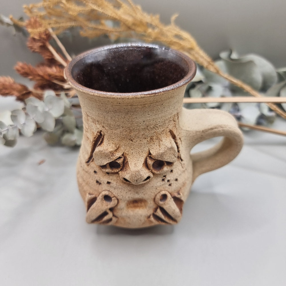 Stoneware face mug with grumpy sculpted expression and rustic brown glaze.