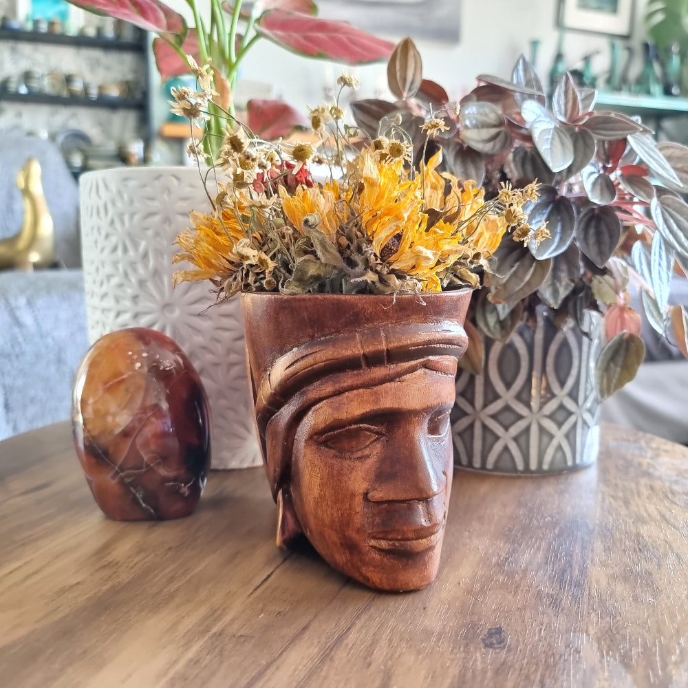 Hand-carved wooden mug featuring a serene face with headdress, styled with dried flowers