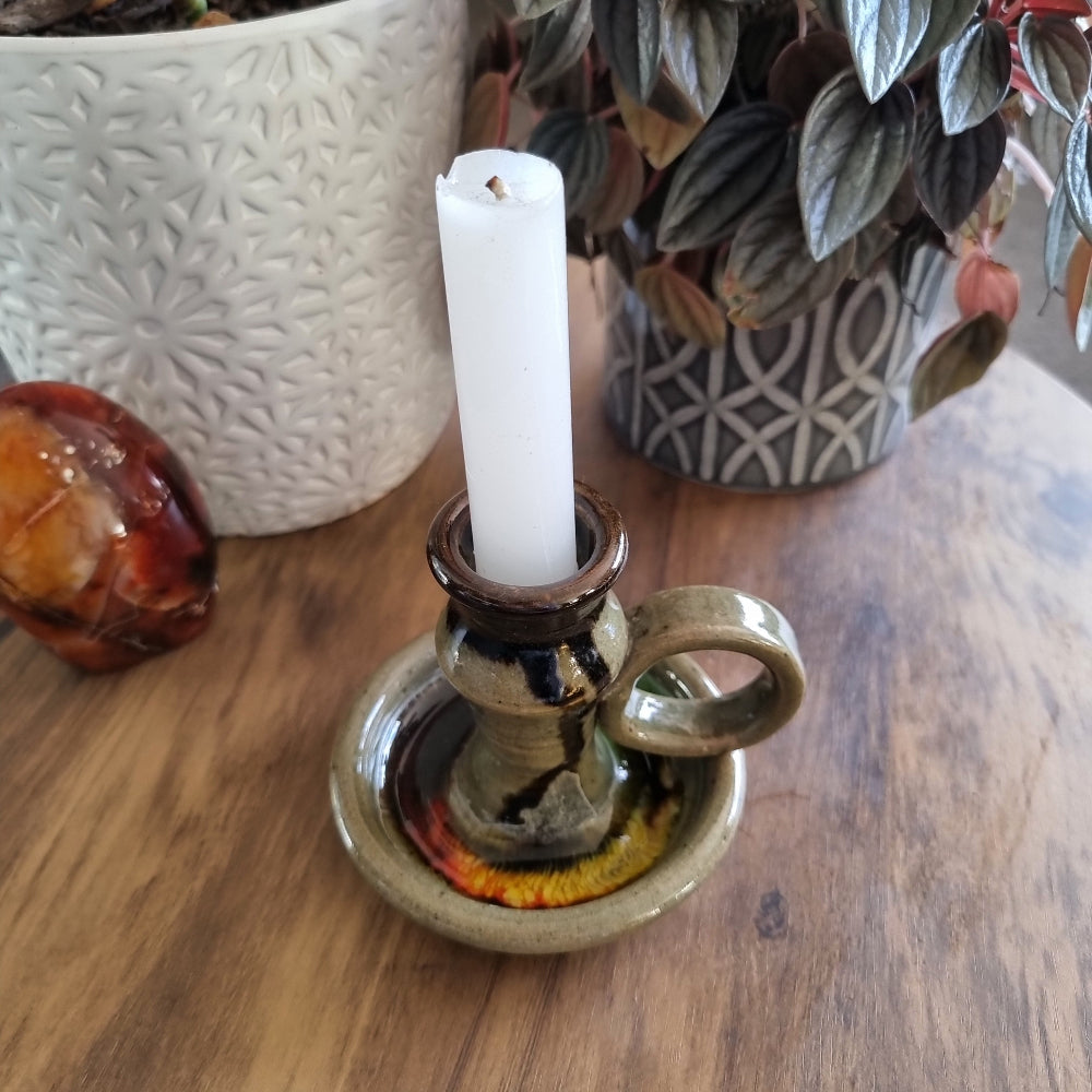 Handmade ceramic chamberstick with earthy glaze and finger loop, holding a white taper candle