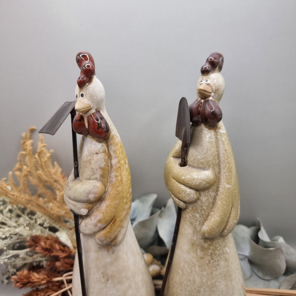 Tall ceramic rooster sculptures holding garden tools, rustic and whimsical style.