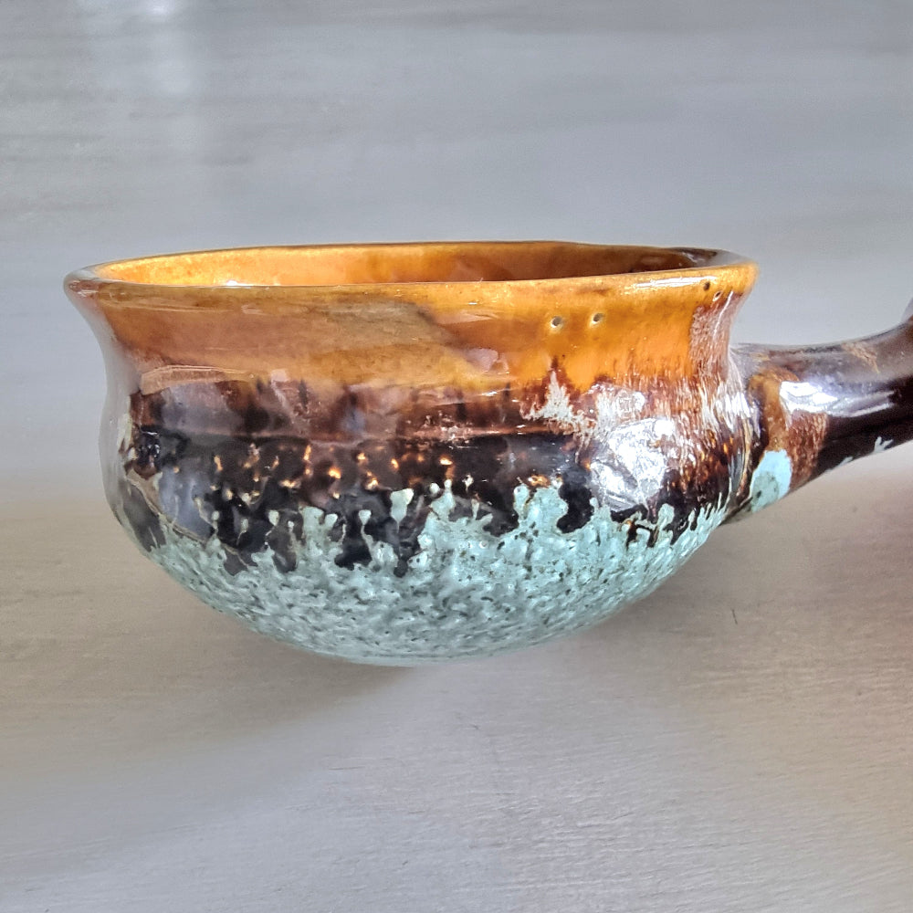Rustic pottery bowl with handle and layered caramel, brown, and green glazes.