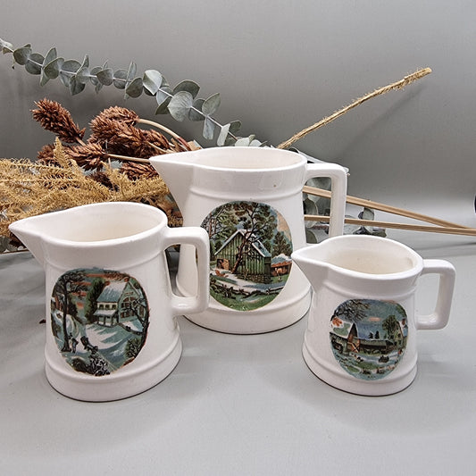 Three white ceramic measuring jugs with colourful winter countryside scenes.
