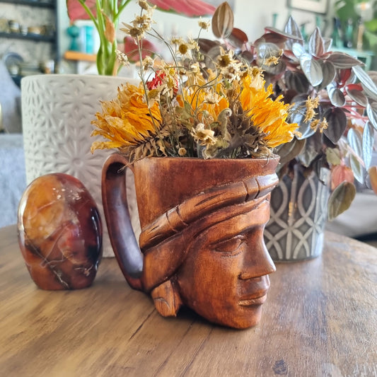 Hand-carved wooden mug featuring a serene face with headdress, styled with dried flowers