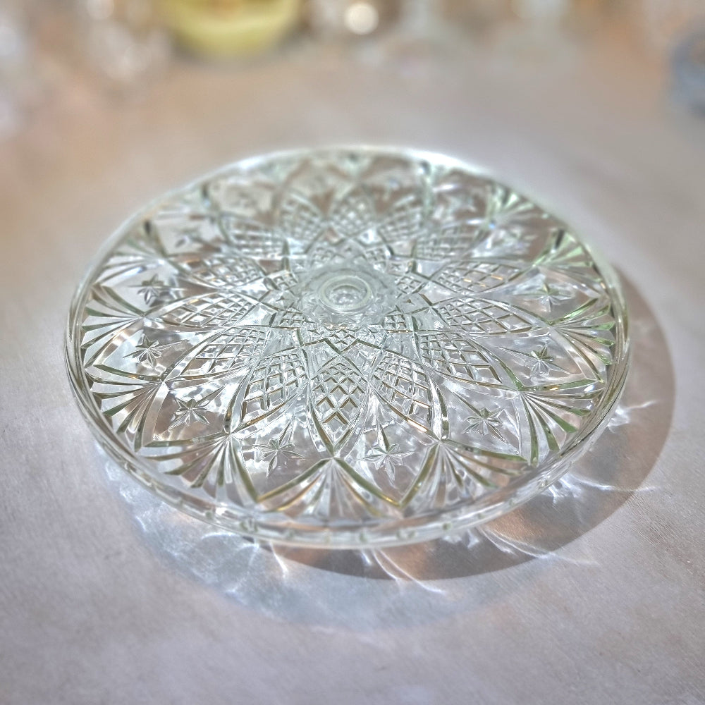 Clear pressed glass cake stand with starburst pattern and raised foot, catching light on a tabletop.