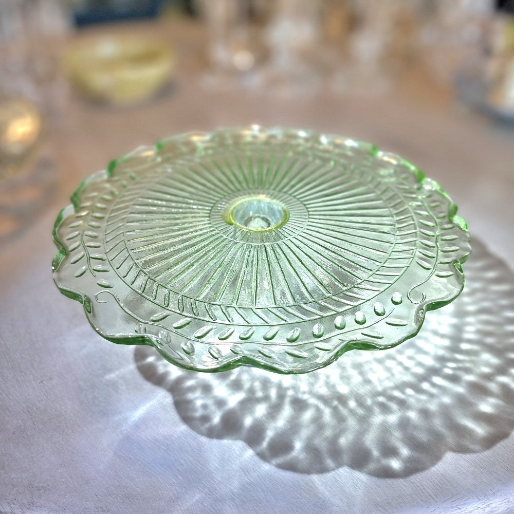 Green pressed glass cake stand with scalloped edge and sunburst pattern.