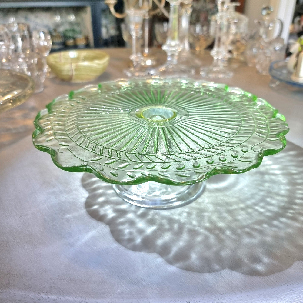 Green pressed glass cake stand with scalloped edge and sunburst pattern.