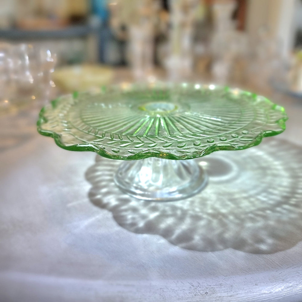 Green pressed glass cake stand with scalloped edge and sunburst pattern.