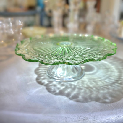 Green pressed glass cake stand with scalloped edge and sunburst pattern.