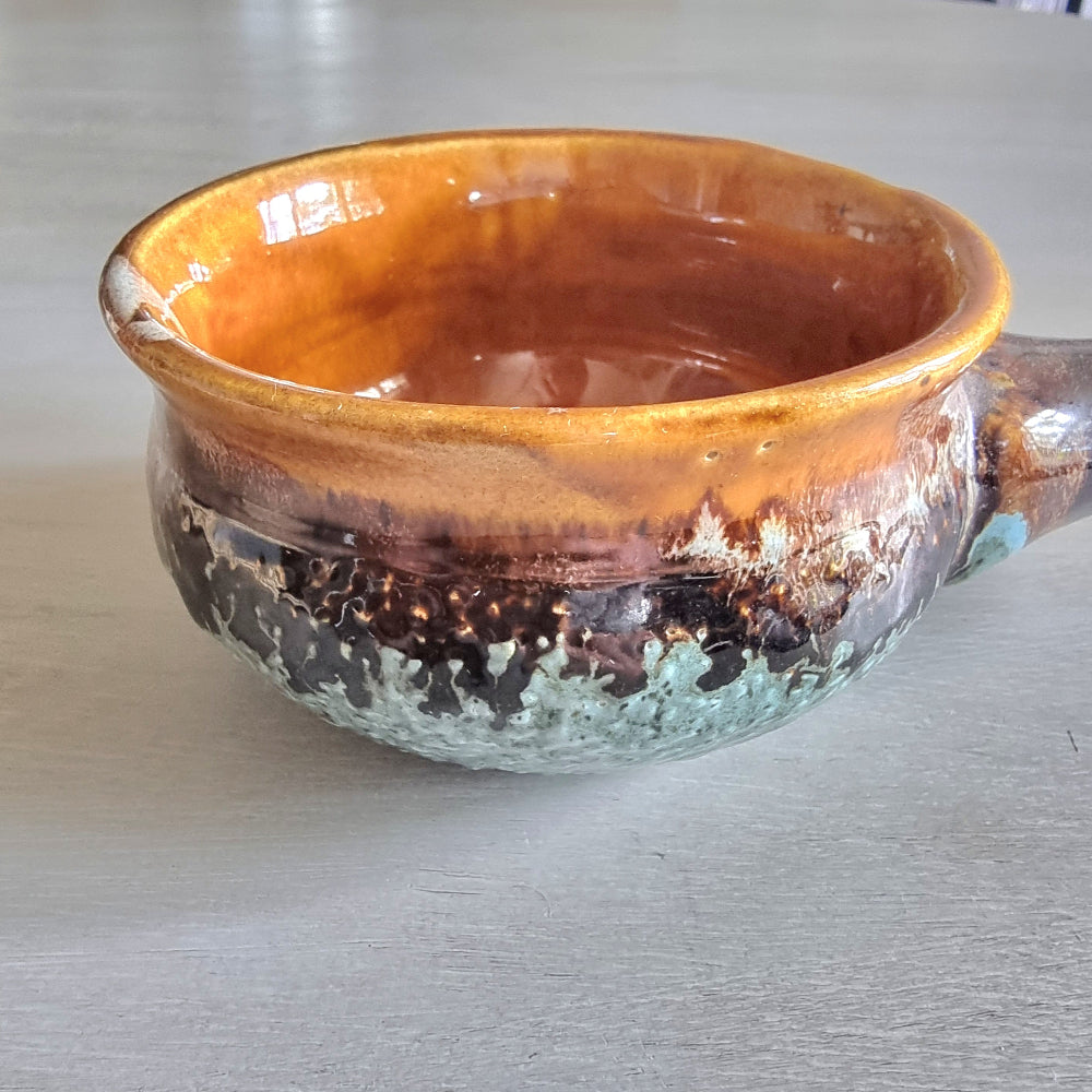 Rustic pottery bowl with handle and layered caramel, brown, and green glazes.