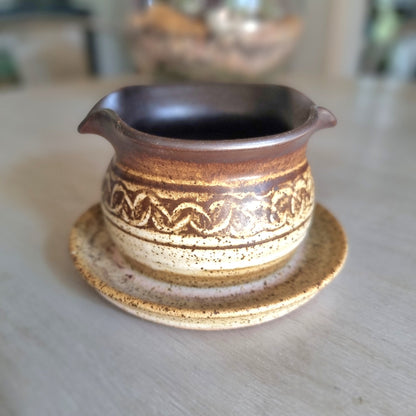 Vintage stoneware gravy jug with brown and cream glaze, speckled finish and pouring spout on a tabletop.
