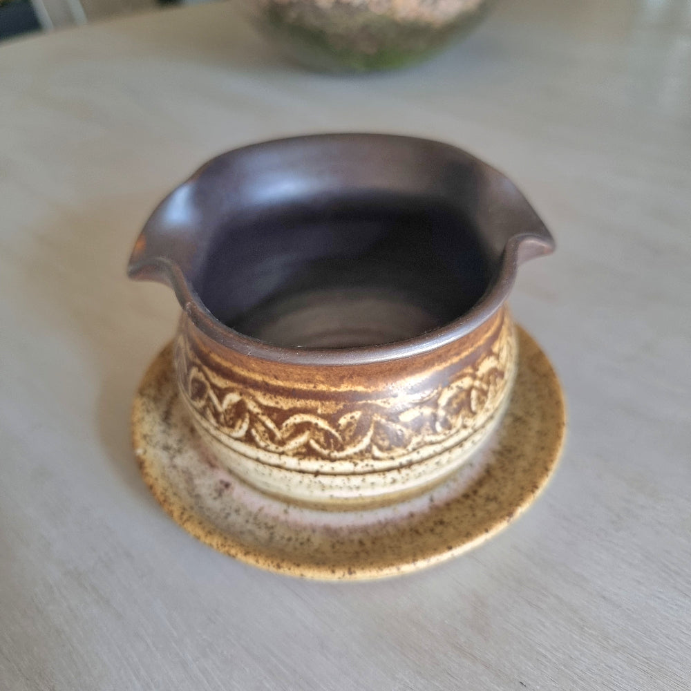 Vintage stoneware gravy jug with brown and cream glaze, speckled finish and pouring spout on a tabletop.