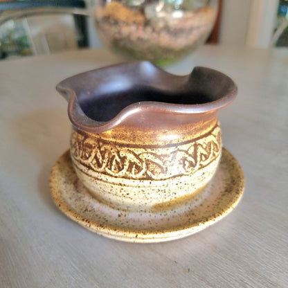 Vintage stoneware gravy jug with brown and cream glaze, speckled finish and pouring spout on a tabletop.