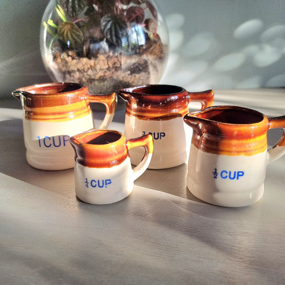 Vintage ceramic measuring jug set with caramel glaze and blue cup markings, including one cracked jug, styled on a table.