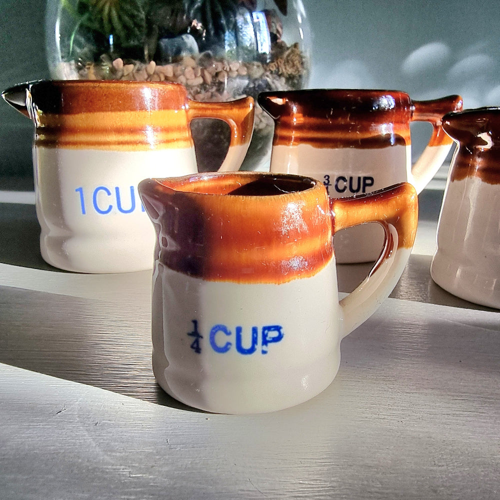 Vintage ceramic measuring jug set with caramel glaze and blue cup markings, including one cracked jug, styled on a table.