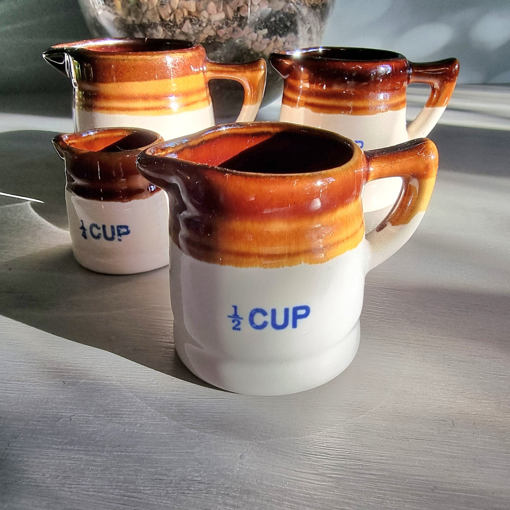 Vintage ceramic measuring jug set with caramel glaze and blue cup markings, including one cracked jug, styled on a table.