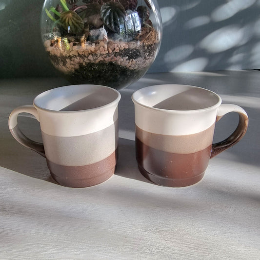 Stoneware mug pair with layered grey and neutral glaze, photographed on a tabletop in natural light.