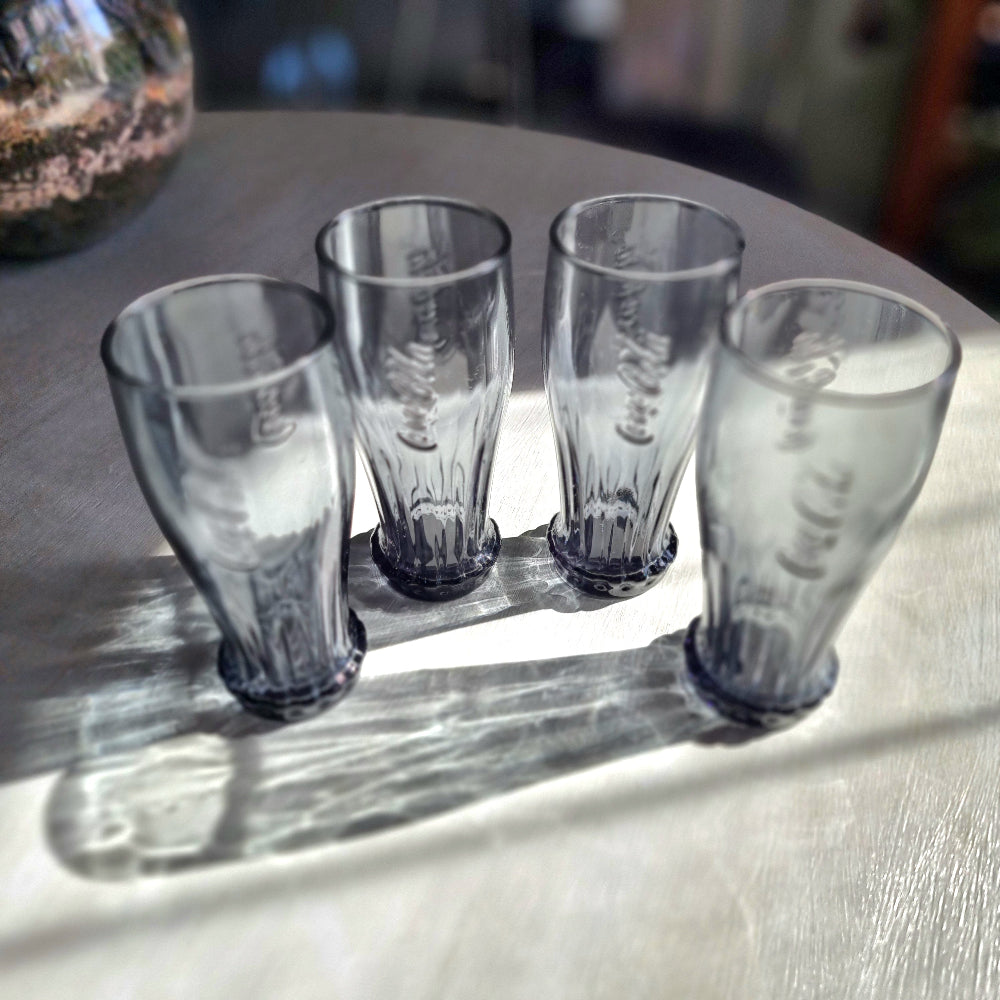 Set of four smoky grey Coca-Cola contour glasses with embossed logo and bottle cap style base.
