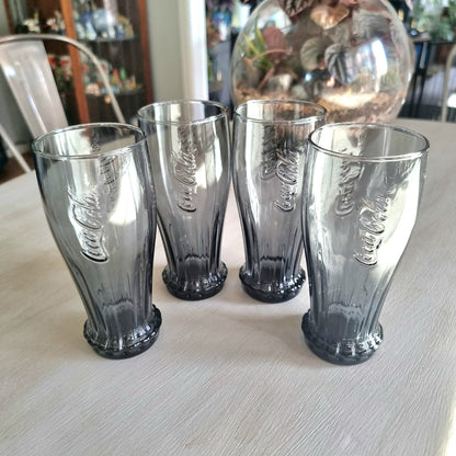 Set of four smoky grey Coca-Cola contour glasses with embossed logo and bottle cap style base.