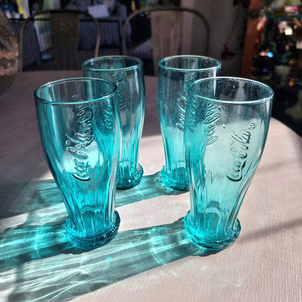 Set of four teal Coca-Cola contour drinking glasses with embossed Coke logo and bottle cap base detail.