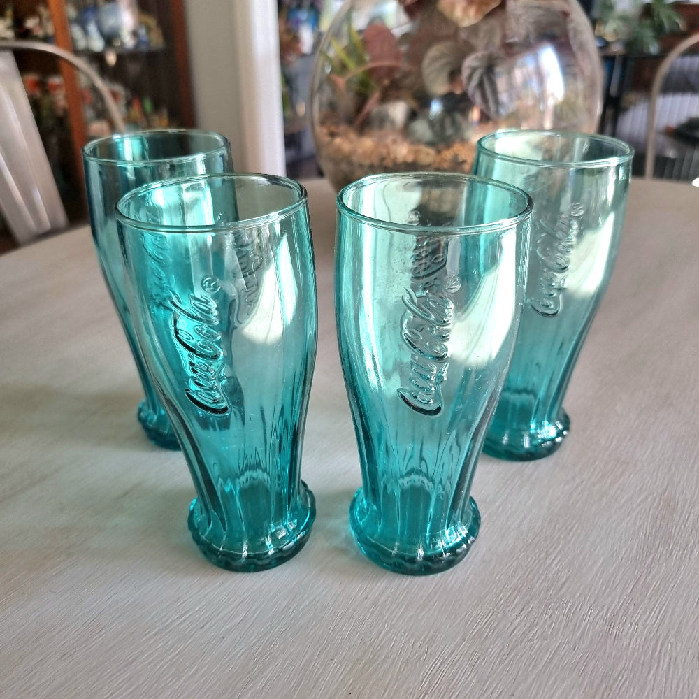Set of four teal Coca-Cola contour drinking glasses with embossed Coke logo and bottle cap base detail.