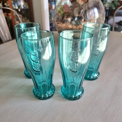 Set of four teal Coca-Cola contour drinking glasses with embossed Coke logo and bottle cap base detail.