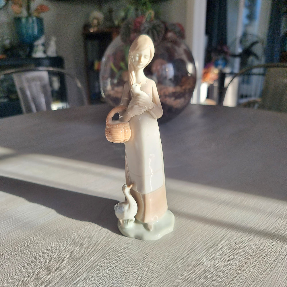 Lladró-inspired porcelain figurine of a young woman with long hair cradling a goose, carrying a basket with a second goose at her feet