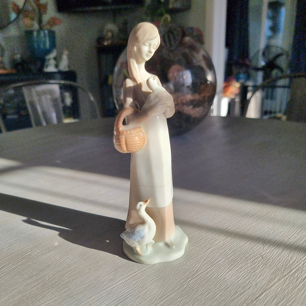 Lladró-inspired porcelain figurine of a young woman with long hair cradling a goose, carrying a basket with a second goose at her feet