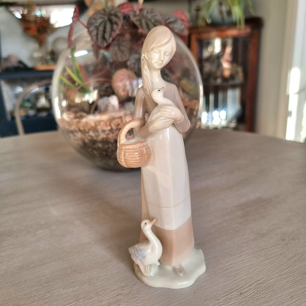 Lladró-inspired porcelain figurine of a young woman with long hair cradling a goose, carrying a basket with a second goose at her feet
