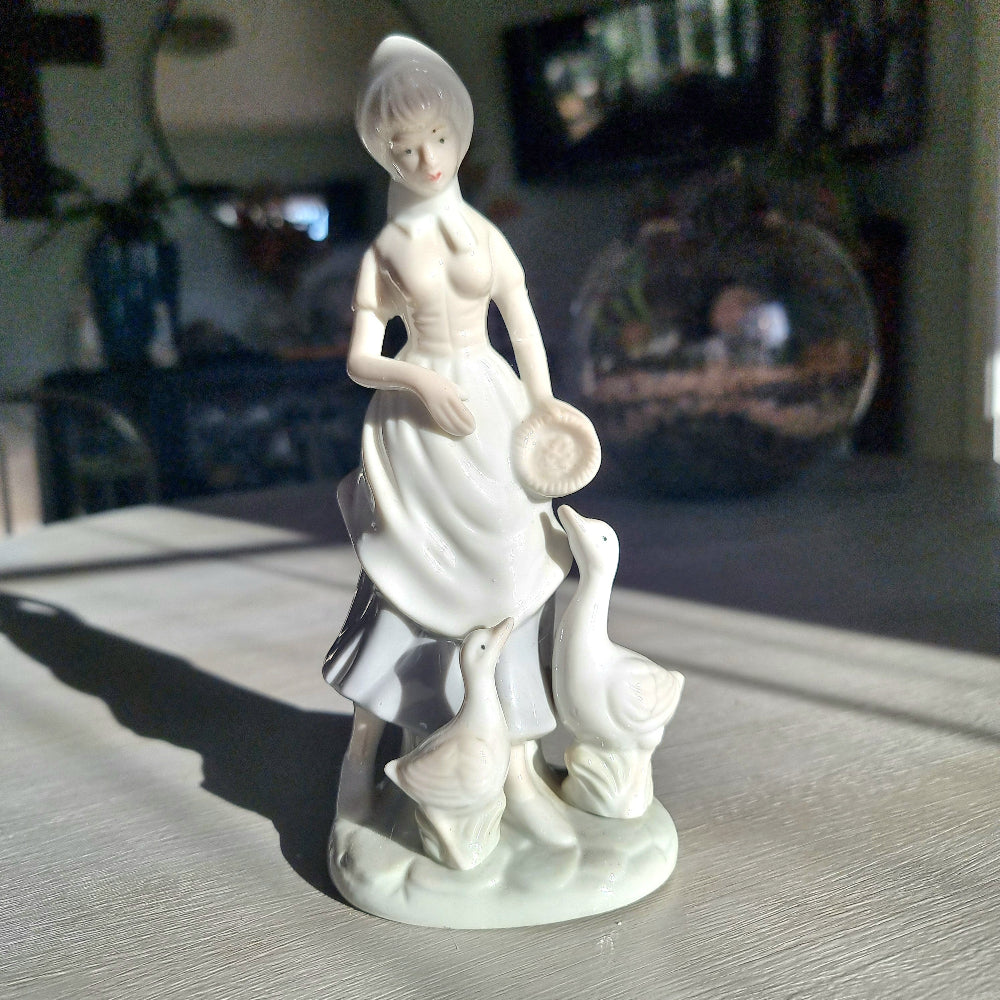 Lladró-inspired porcelain figurine of a country girl in bonnet and apron holding a dish, with two white geese at her feet