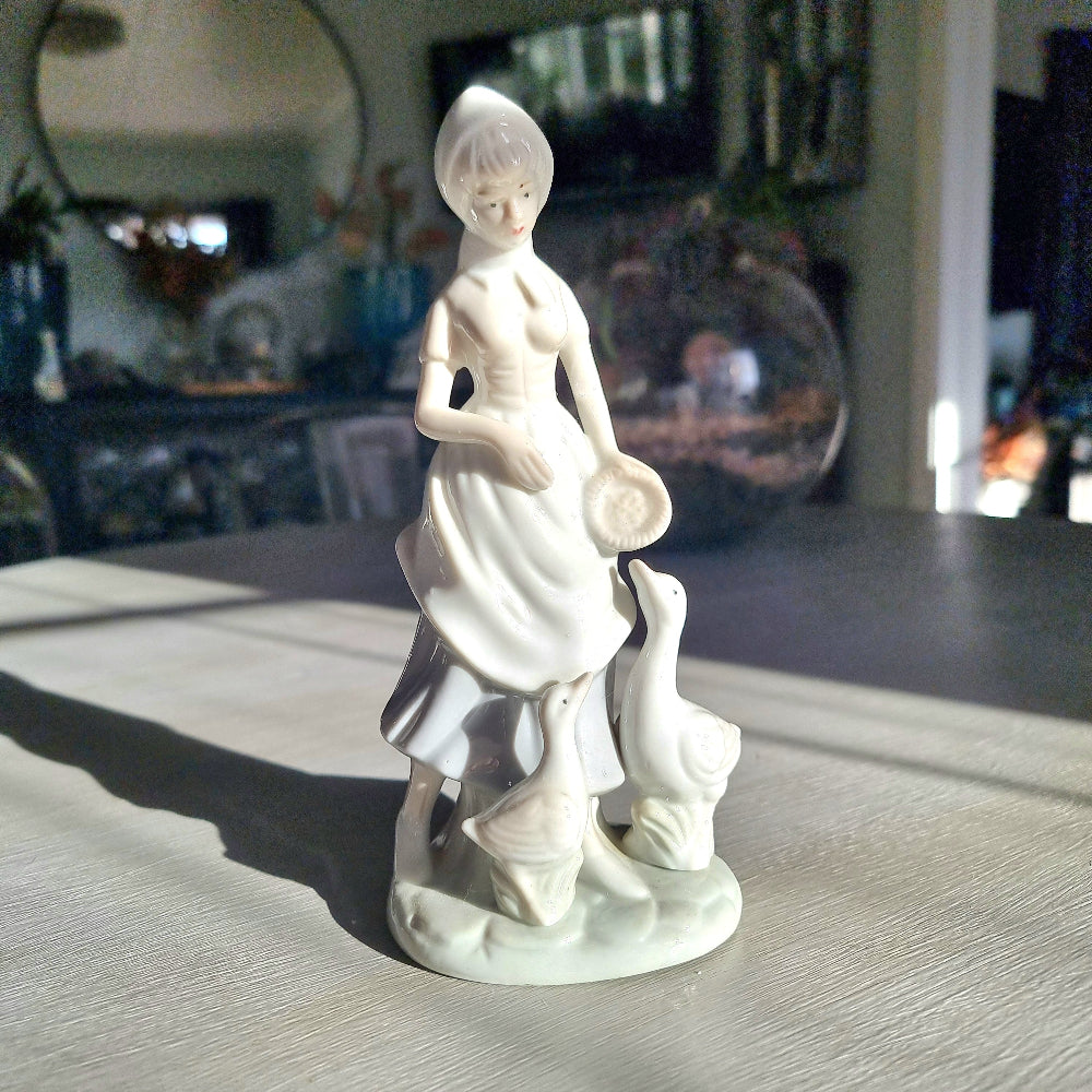 Lladró-inspired porcelain figurine of a country girl in bonnet and apron holding a dish, with two white geese at her feet