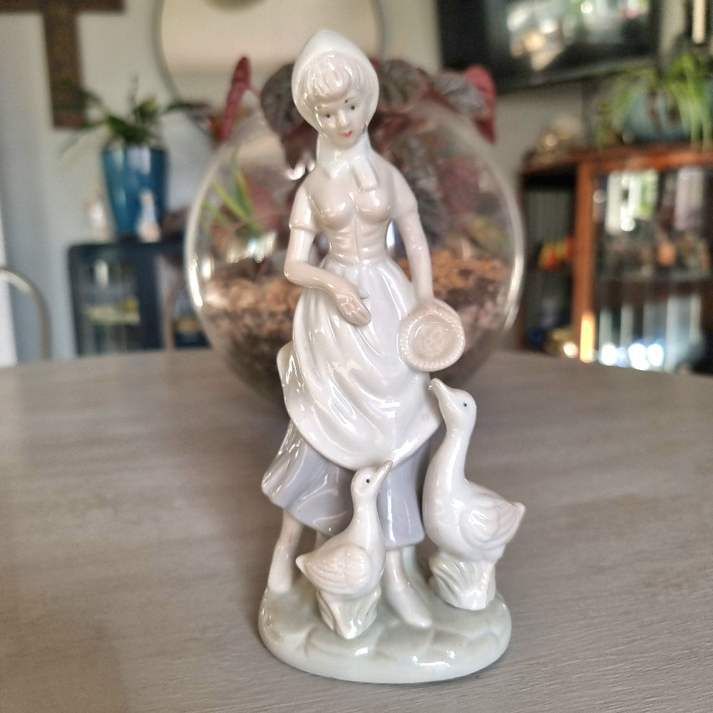 Lladró-inspired porcelain figurine of a country girl in bonnet and apron holding a dish, with two white geese at her feet