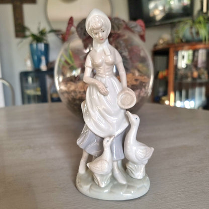 Lladró-inspired porcelain figurine of a country girl in bonnet and apron holding a dish, with two white geese at her feet