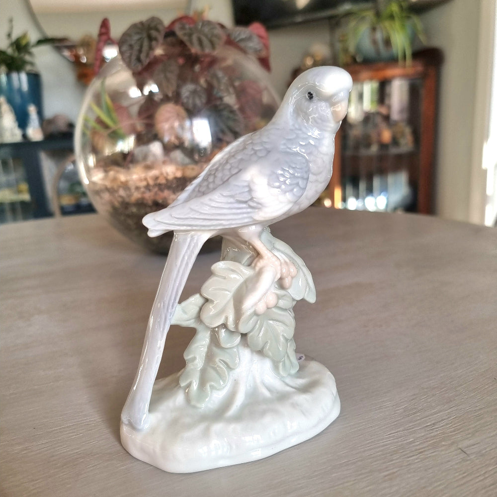 Vintage porcelain budgerigar figurine in glossy white glaze perched on a branch with sculpted leaf base