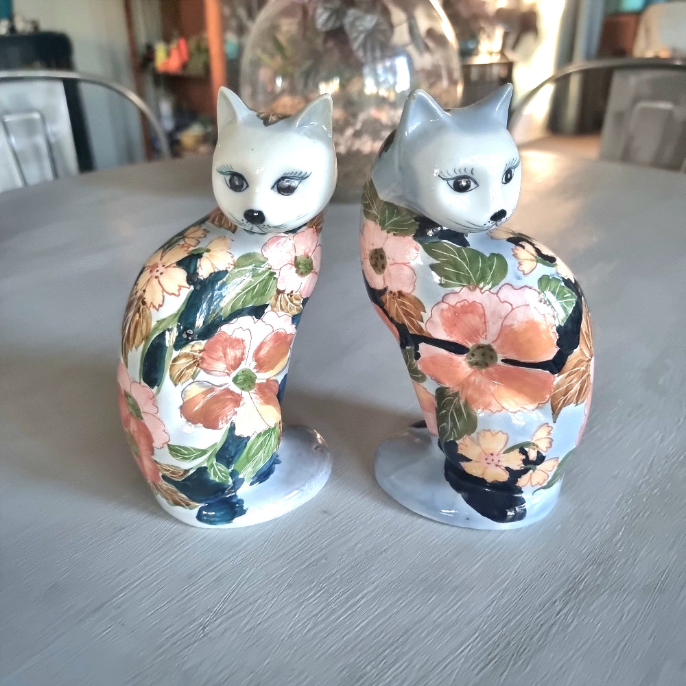 Pair of vintage hand-painted ceramic cat figurines with pink coral and yellow floral design on white and periwinkle glaze, seated upright with painted face and dark expressive eyes