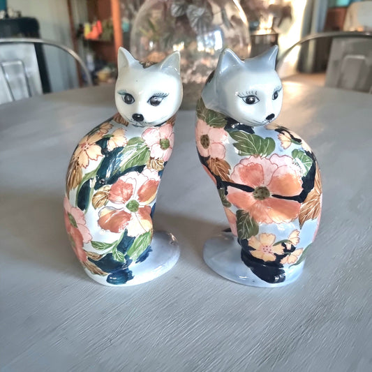 Pair of vintage hand-painted ceramic cat figurines with pink coral and yellow floral design on white and periwinkle glaze, seated upright with painted face and dark expressive eyes