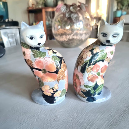 Pair of vintage hand-painted ceramic cat figurines with pink coral and yellow floral design on white and periwinkle glaze, seated upright with painted face and dark expressive eyes