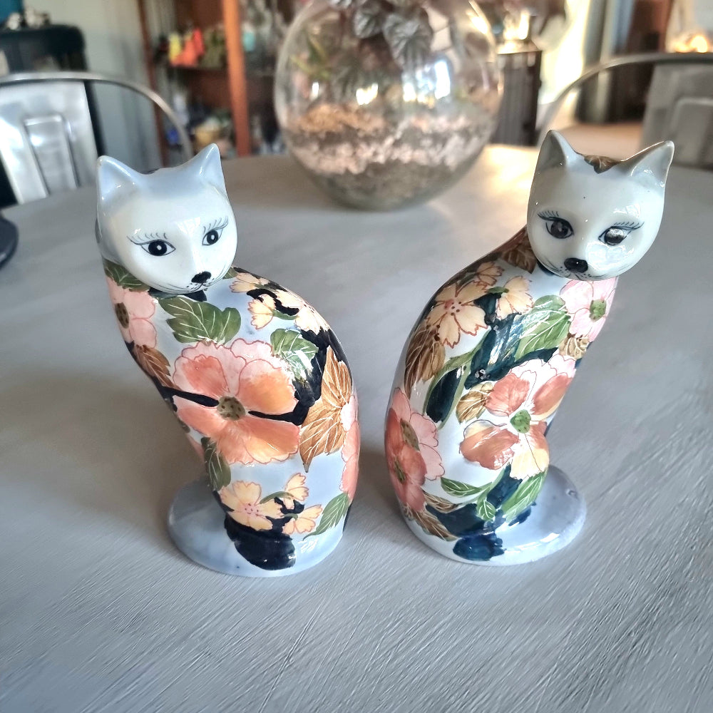 Pair of vintage hand-painted ceramic cat figurines with pink coral and yellow floral design on white and periwinkle glaze, seated upright with painted face and dark expressive eyes
