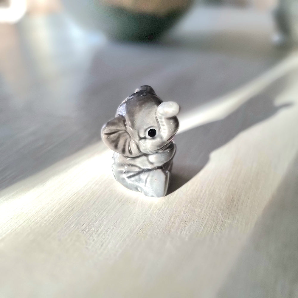 Small grey ceramic elephant figurine sitting with trunk raised, viewed from the front, on a light wooden surface