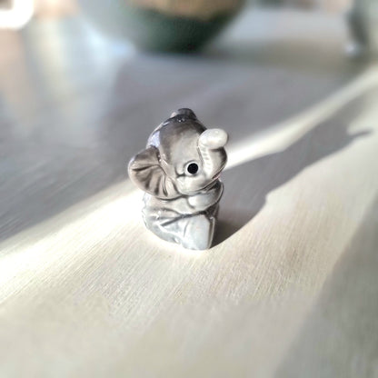 Small grey ceramic elephant figurine sitting with trunk raised, viewed from the front, on a light wooden surface