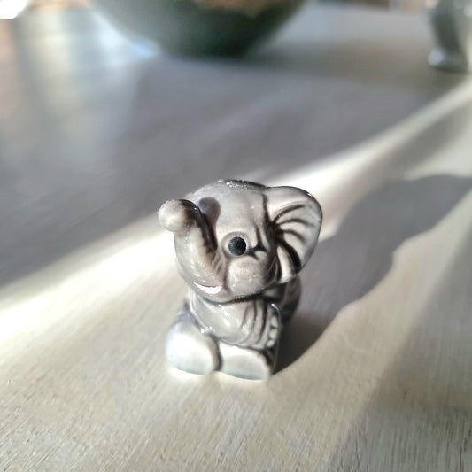 Small grey ceramic elephant figurine sitting with trunk raised, viewed from the front, on a light wooden surface
