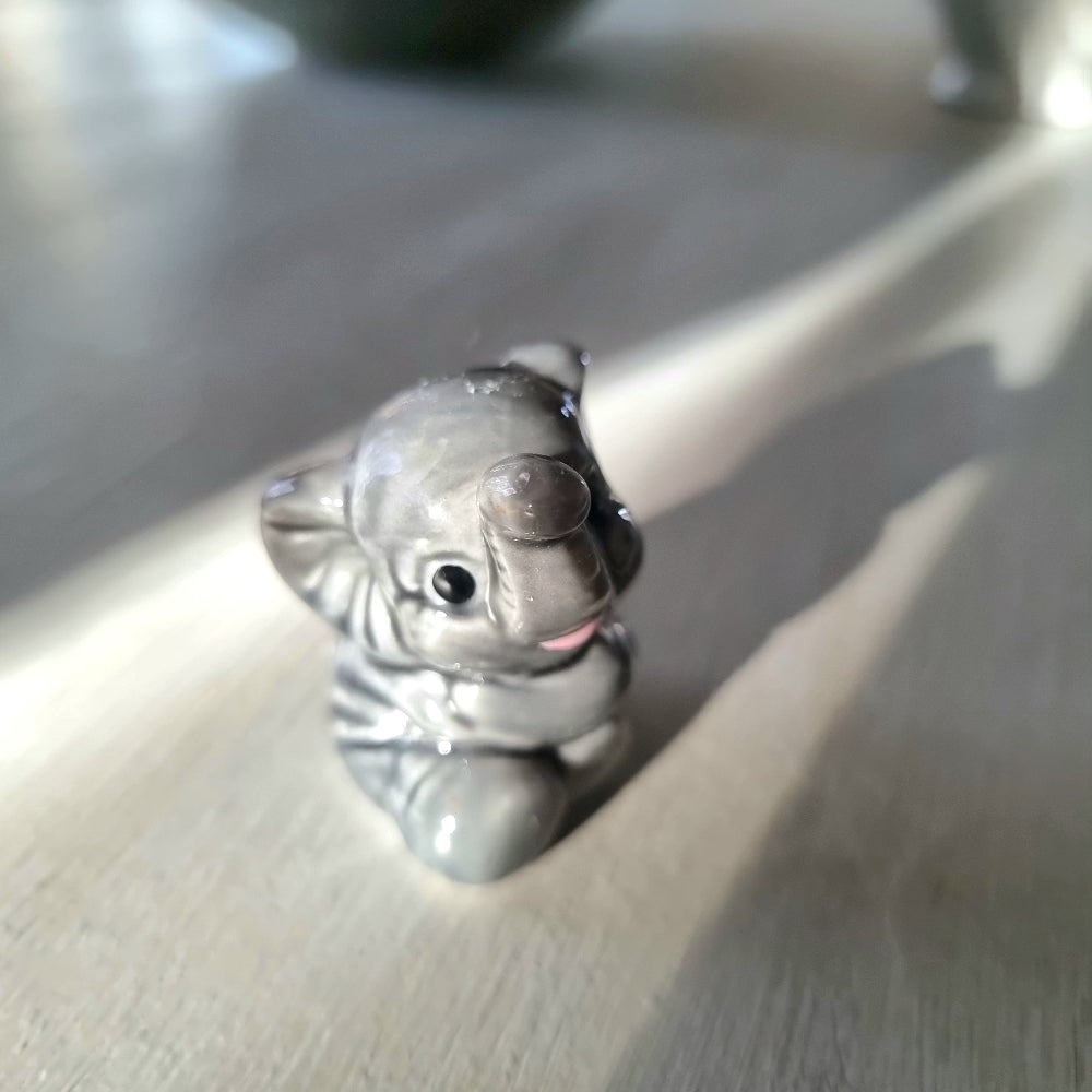 Small grey ceramic elephant figurine sitting with trunk raised, viewed from the front, on a light wooden surface
