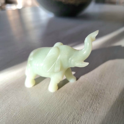 Small carved pale green stone elephant figurine with raised trunk, standing pose, on a light wooden surface in natural light