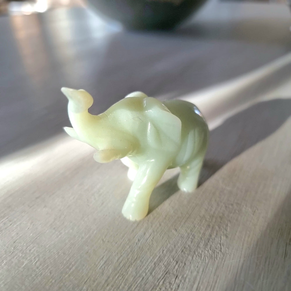 Small carved pale green stone elephant figurine with raised trunk, standing pose, on a light wooden surface in natural light