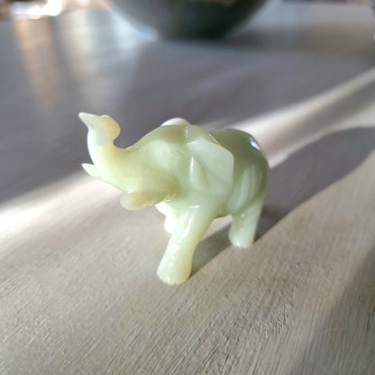 Small carved pale green stone elephant figurine with raised trunk, standing pose, on a light wooden surface in natural light