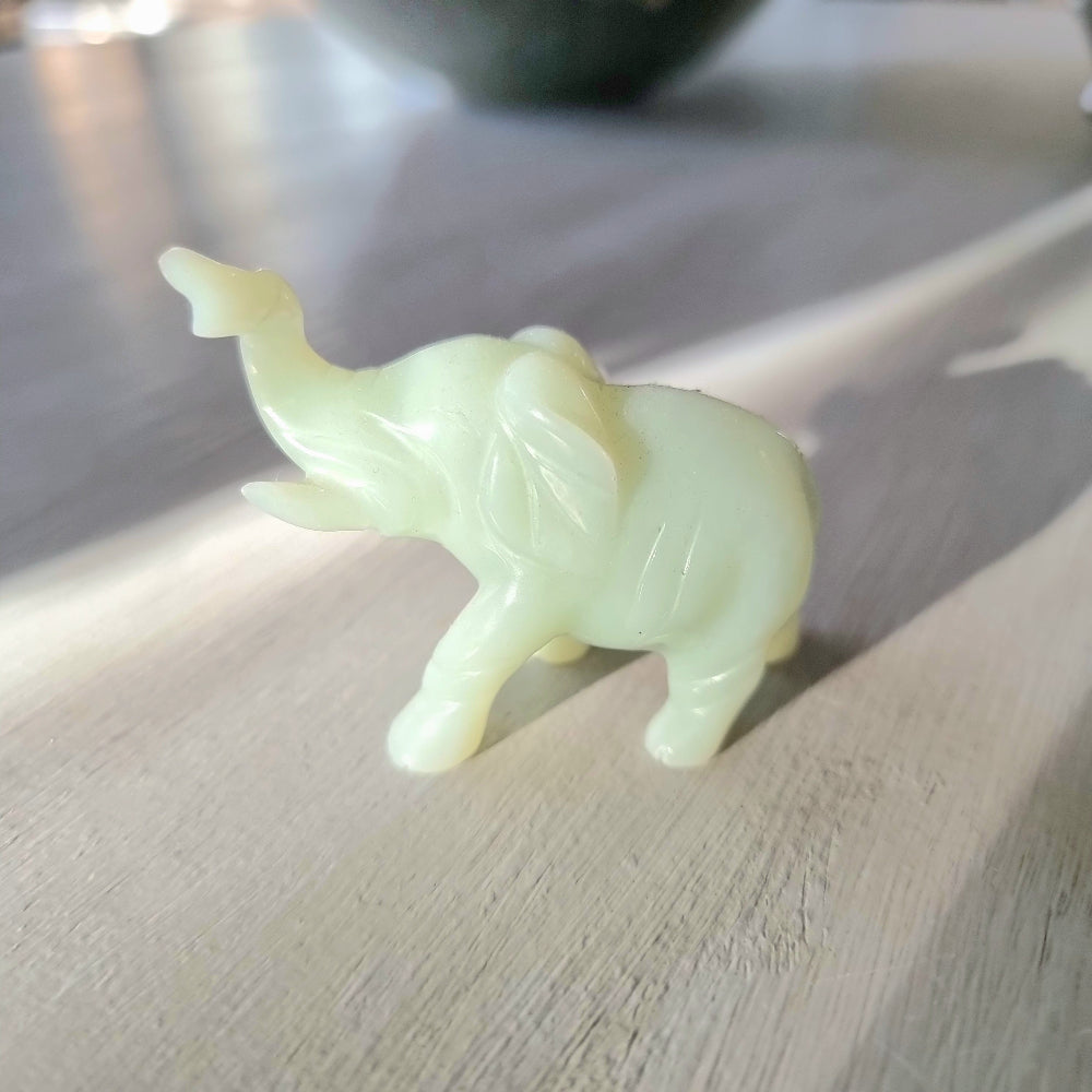 Small carved pale green stone elephant figurine with raised trunk, standing pose, on a light wooden surface in natural light