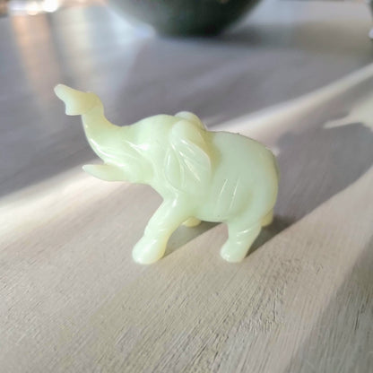 Small carved pale green stone elephant figurine with raised trunk, standing pose, on a light wooden surface in natural light